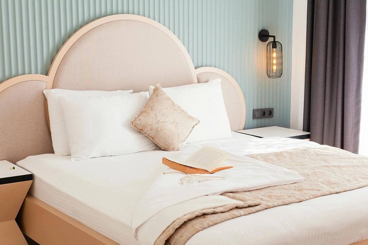 Neatly made bed with white linens, a beige throw, a decorative pillow, an open book, and soft lighting beside a pastel green wall.