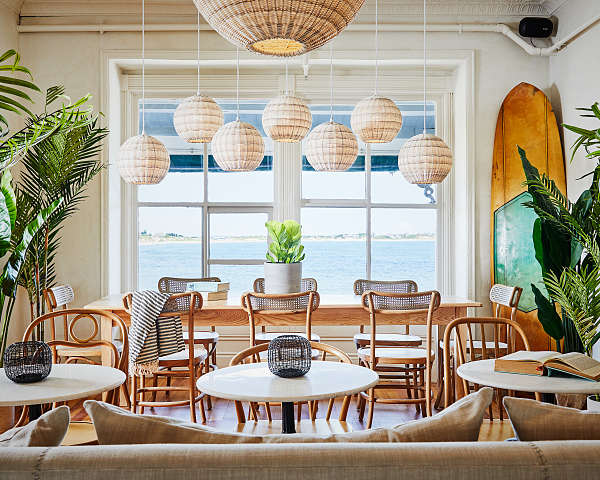 Cozy cafe with rattan chairs, hanging wicker lamps, potted plants, and large windows overlooking a serene ocean view.