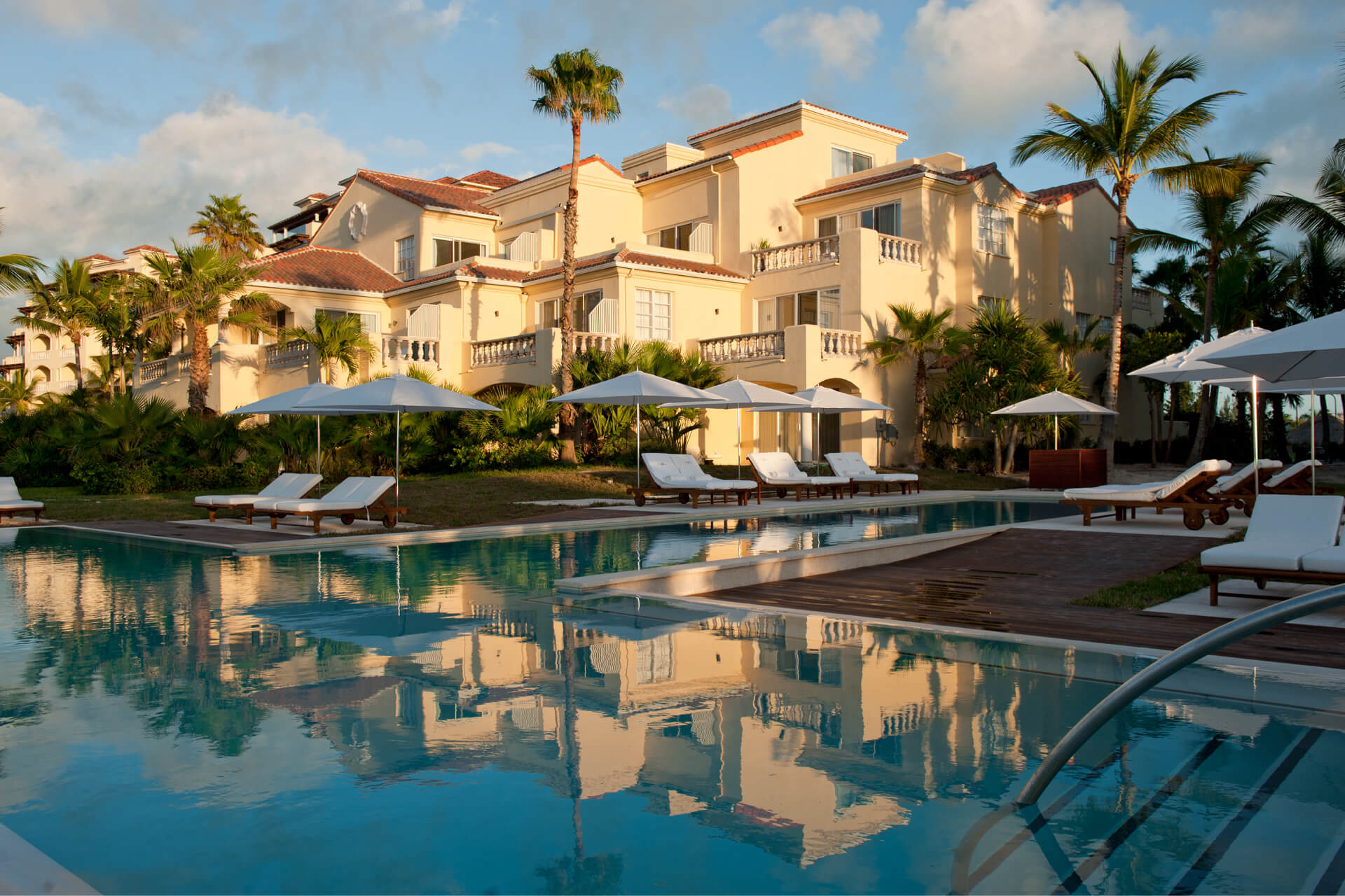 Luxury resort with cream-colored buildings, palm trees, lounge chairs, and umbrellas around a clear swimming pool reflecting the scenery at sunset.