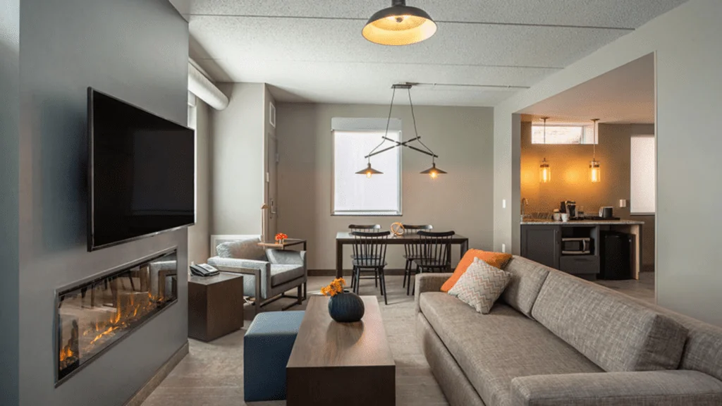 Modern living room with gray sofa, mounted TV, fireplace, dining area, and kitchen. Warm lighting and minimalist decor create a cozy atmosphere.