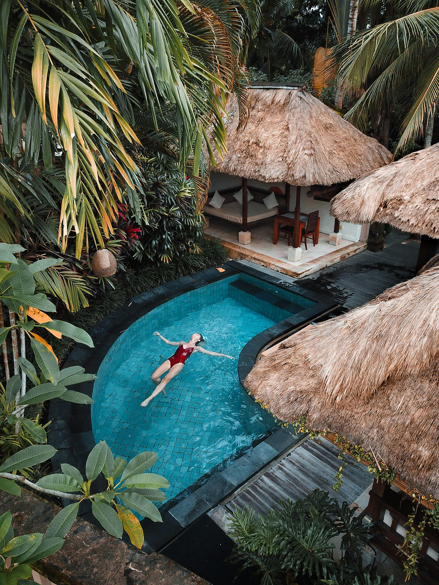 A person in a red swimsuit floats in a turquoise pool surrounded by tropical plants and thatched-roof huts.