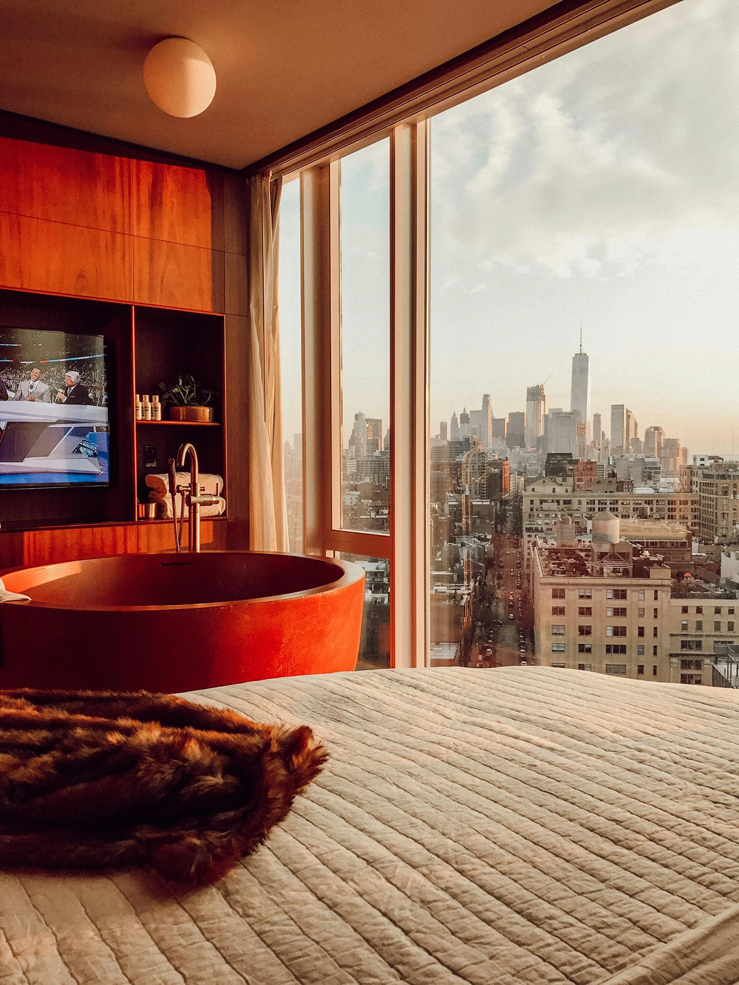 Modern hotel room with a red bathtub, TV, and large window overlooking a sunlit city skyline with tall buildings.