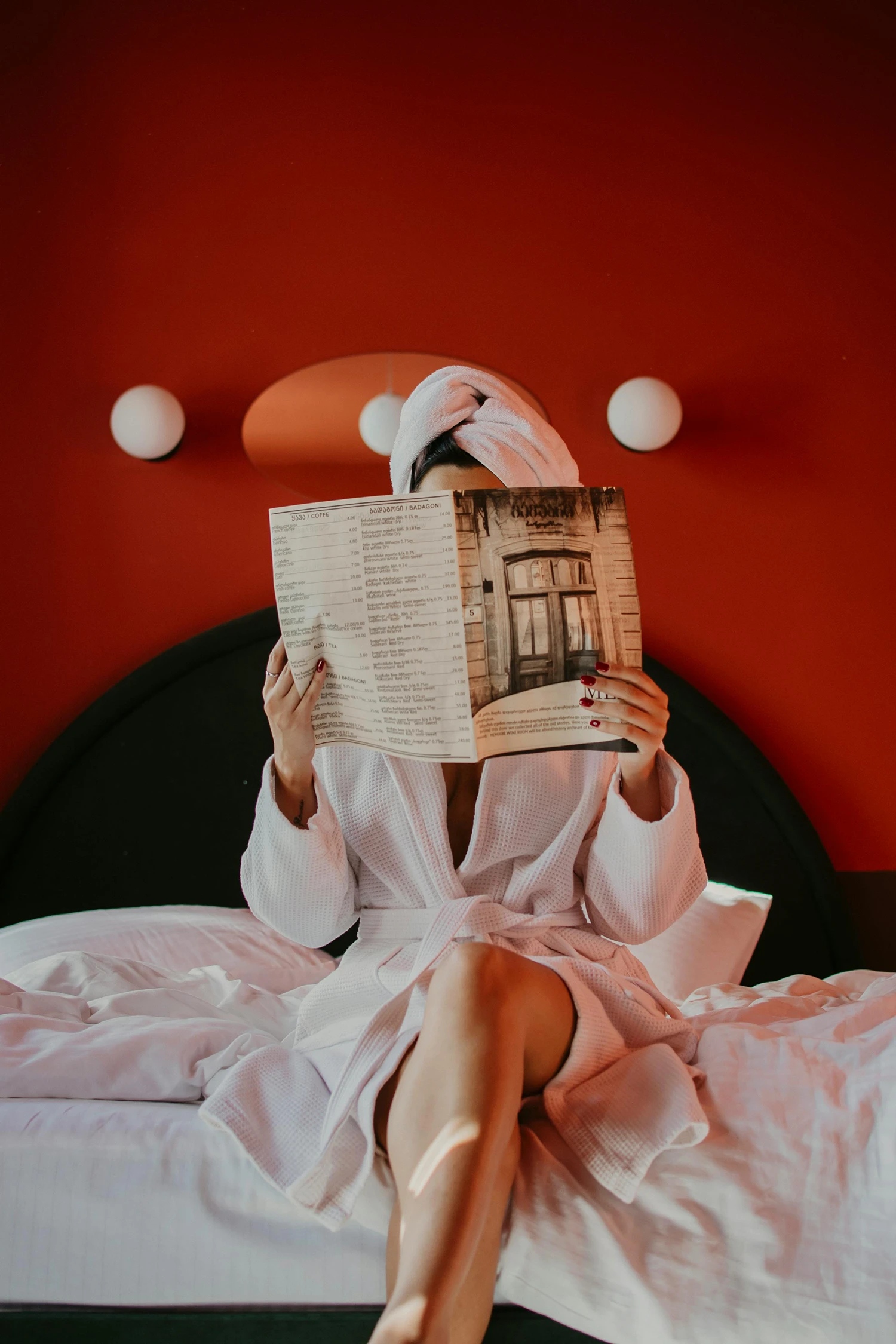 A person in a bathrobe and towel sits on a bed with white sheets, reading a magazine against a red wall with round white lights.
