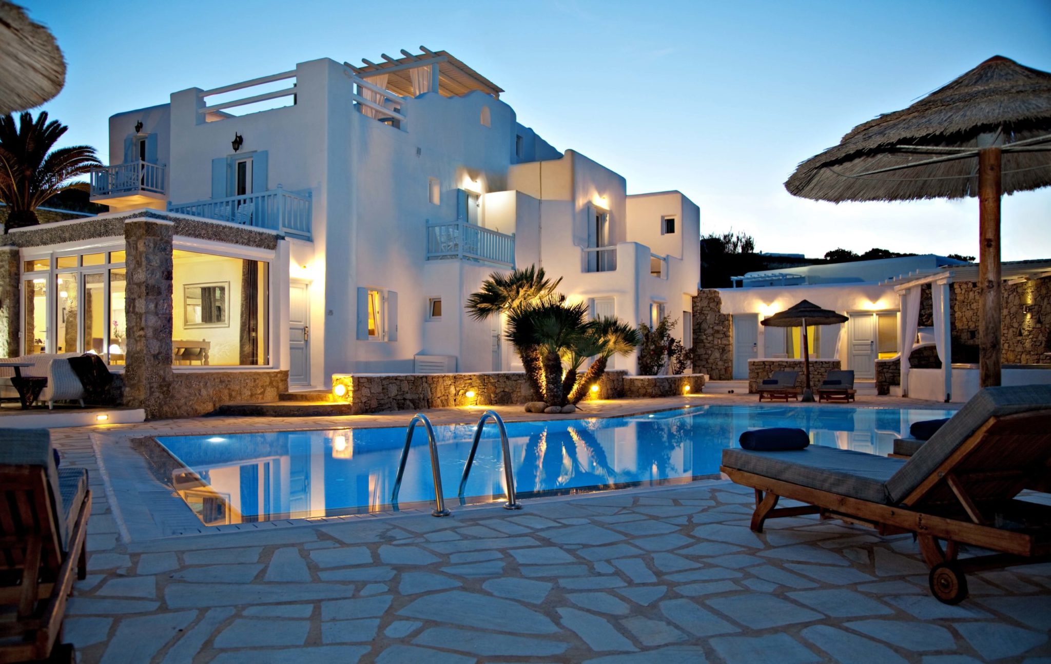 Luxury white villa with balconies, palm trees, and an illuminated swimming pool with lounge chairs and umbrellas at dusk.