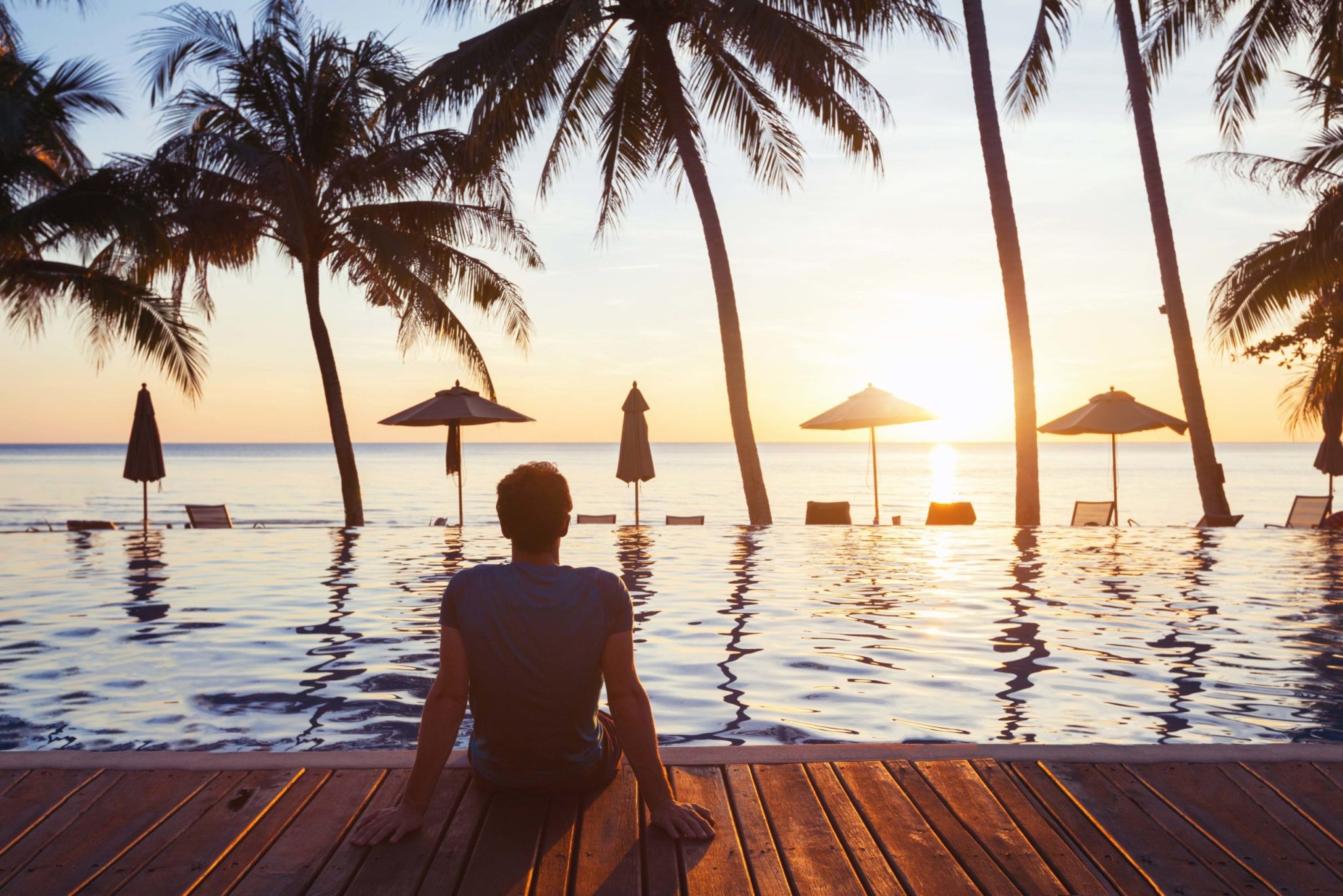 A person sits by a pool facing palm trees and the ocean, watching the sunset. Beach umbrellas and lounge chairs line the water’s edge.