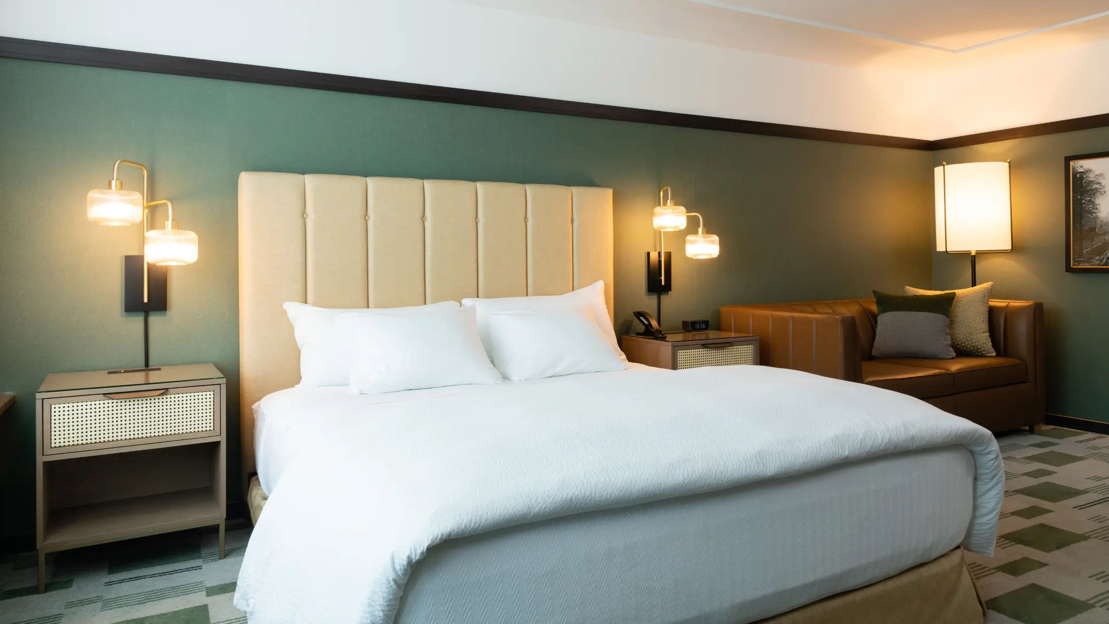 Modern hotel room with a large bed, beige headboard, bedside tables, two wall lamps, a brown sofa, and a standing floor lamp.