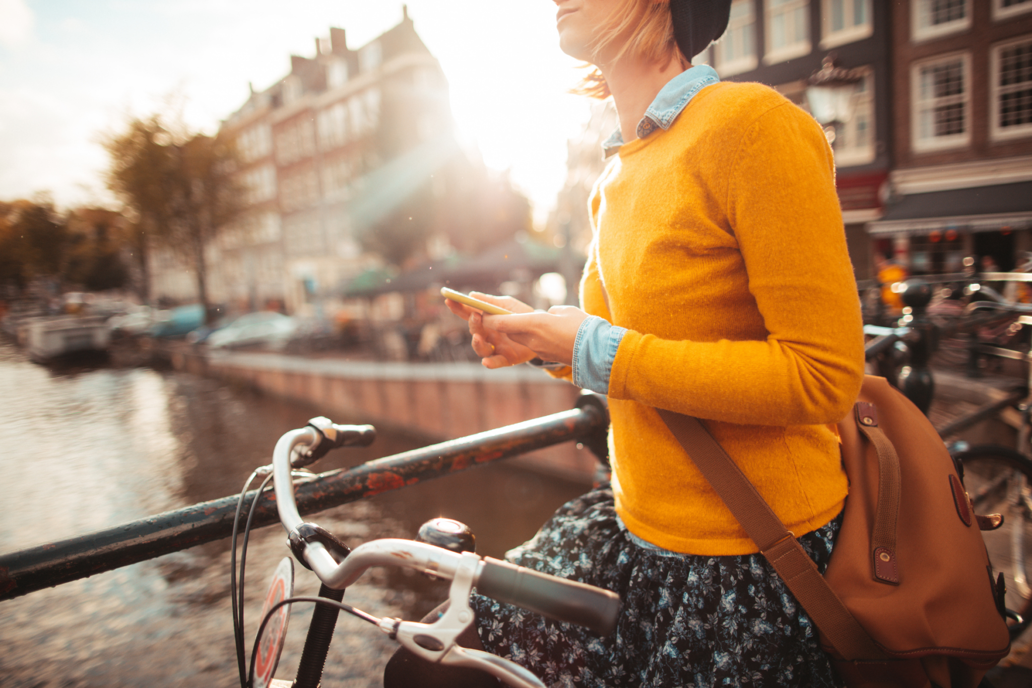 Person in a yellow sweater with a backpack stands by a bicycle on a canal bridge, using a phone in a sunny city setting.