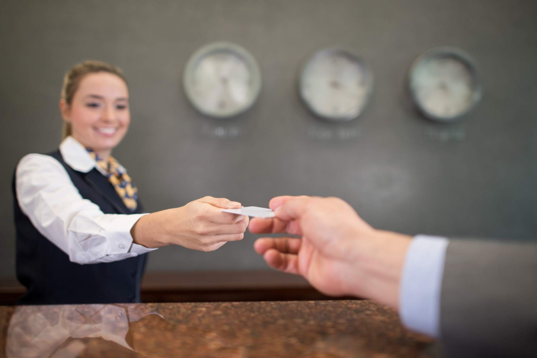 A hotel receptionist hands a key card to a guest across the front desk, with clocks showing different time zones in the background.