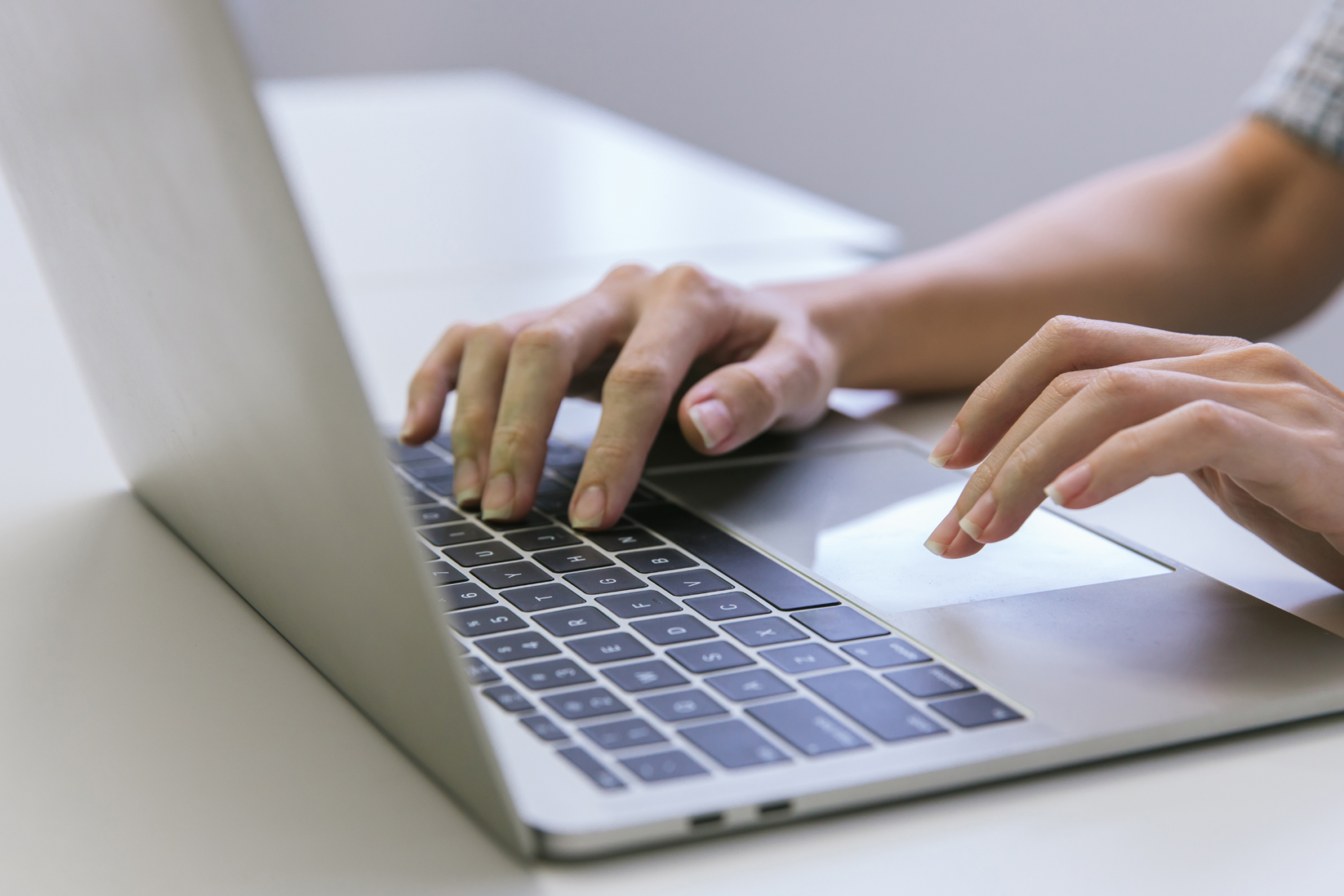 Close-up of a person’s hands typing on a laptop keyboard at a white desk, with natural light in the background.