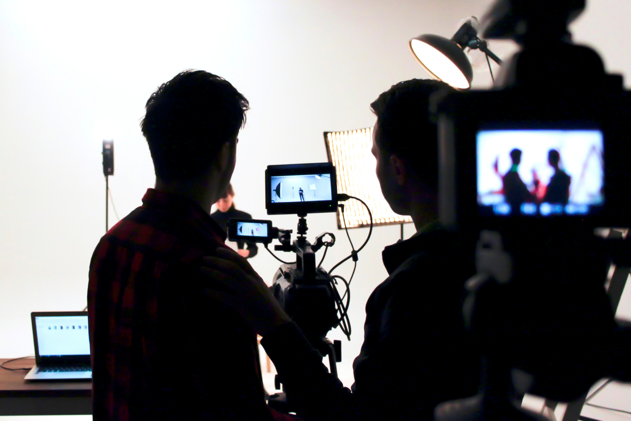 Two people operate a camera on a film set, checking footage on screens with studio lights and equipment visible in the background.