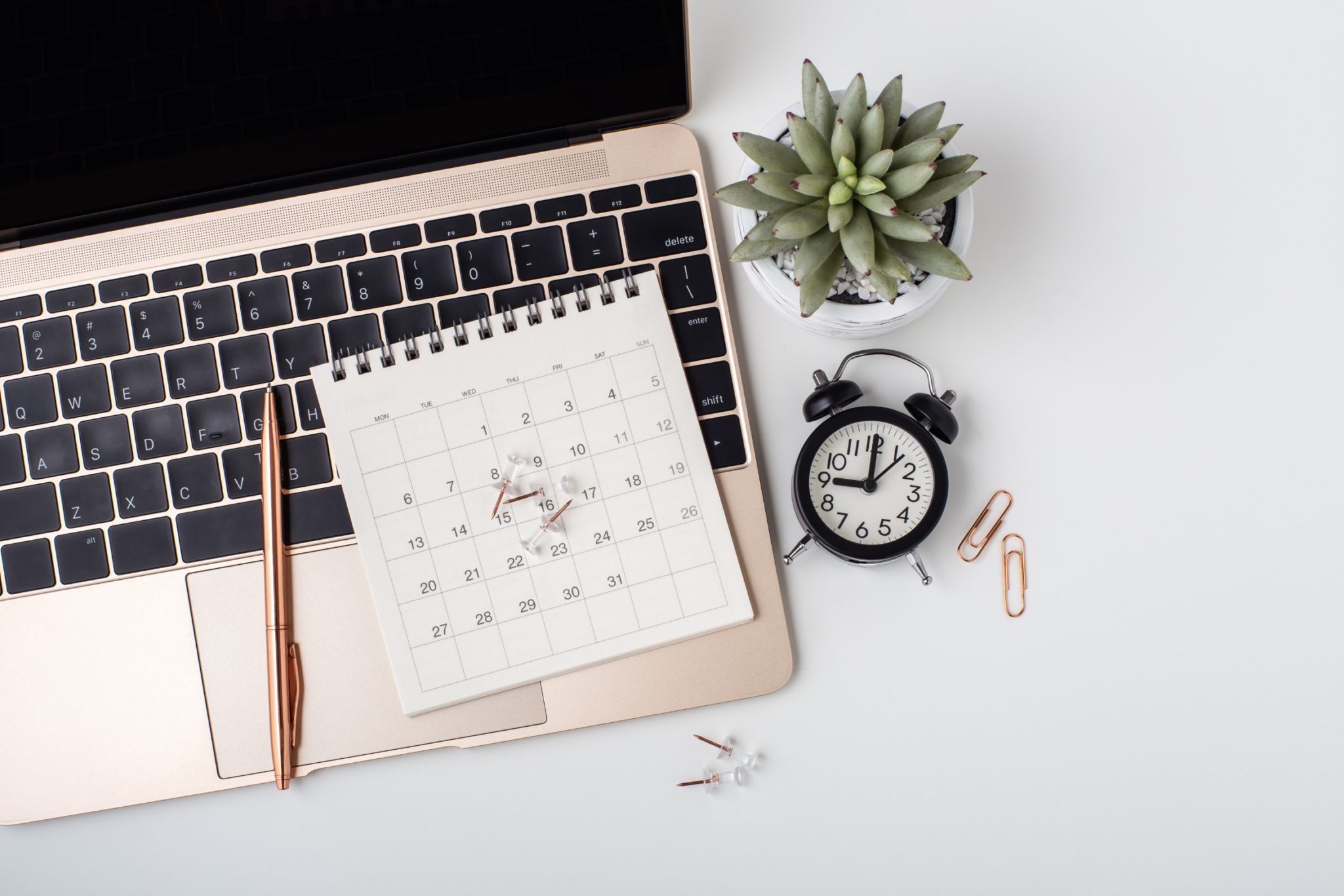 A calendar, pen, and push pins on a laptop keyboard, next to a small potted succulent, an alarm clock, and paper clips on a white desk.