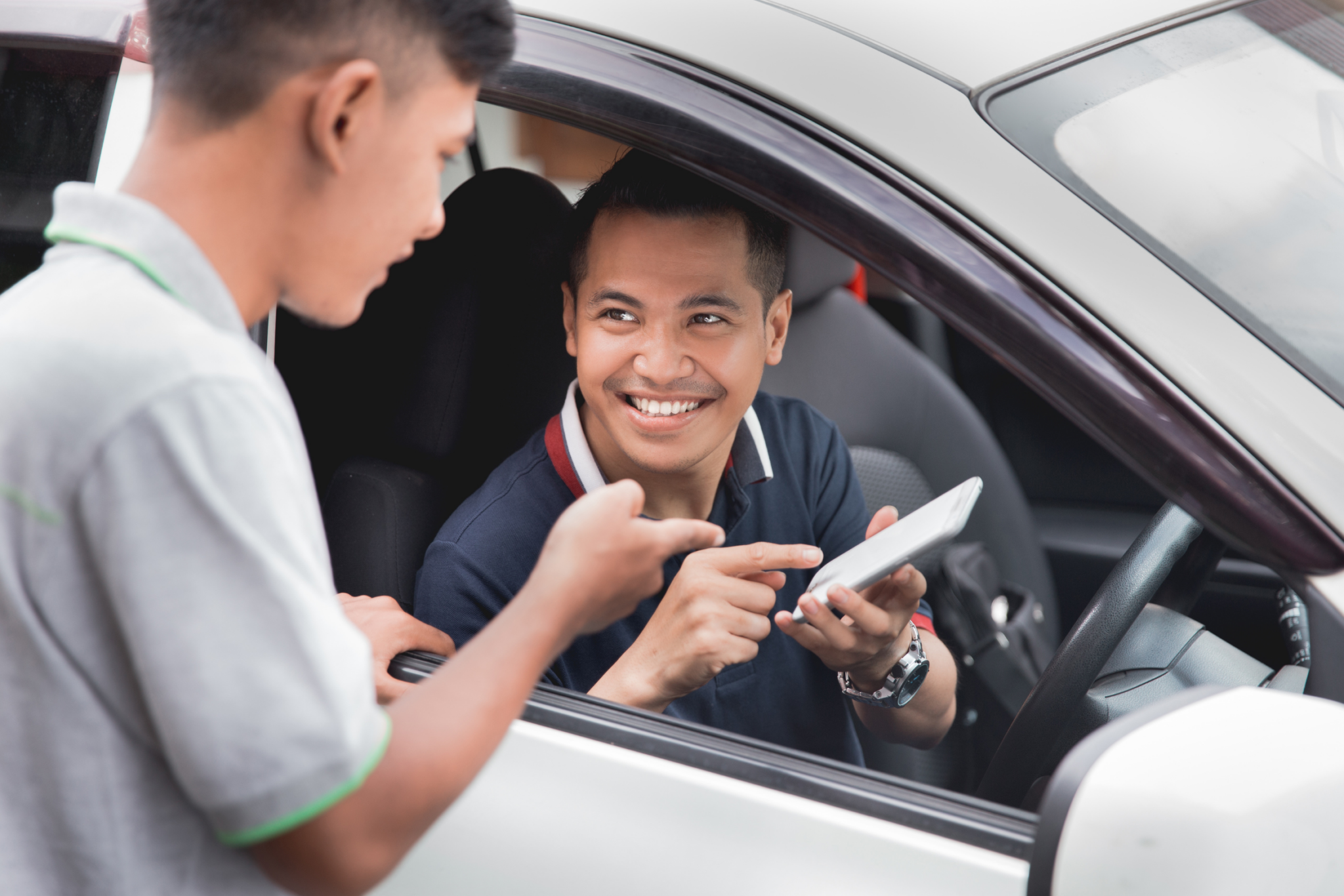 Smiling man in a car points at a tablet while talking to another man standing outside the drivers window.
